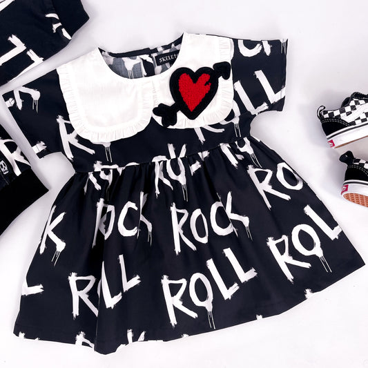 Kids black smock dress with a large frilled, heart detailed collar and the words "rock" and "roll" printed on it repeatedly