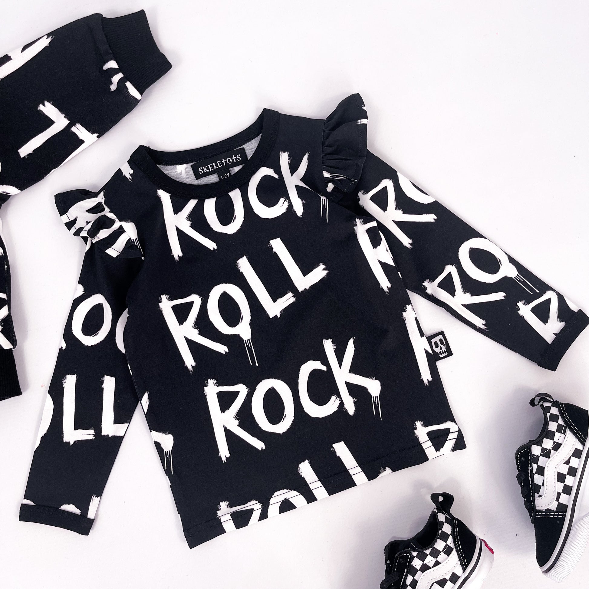 Kids black long sleeved sweatshirt with frilled shoulder detail and words "rock" and "roll" printed on it repeatedly